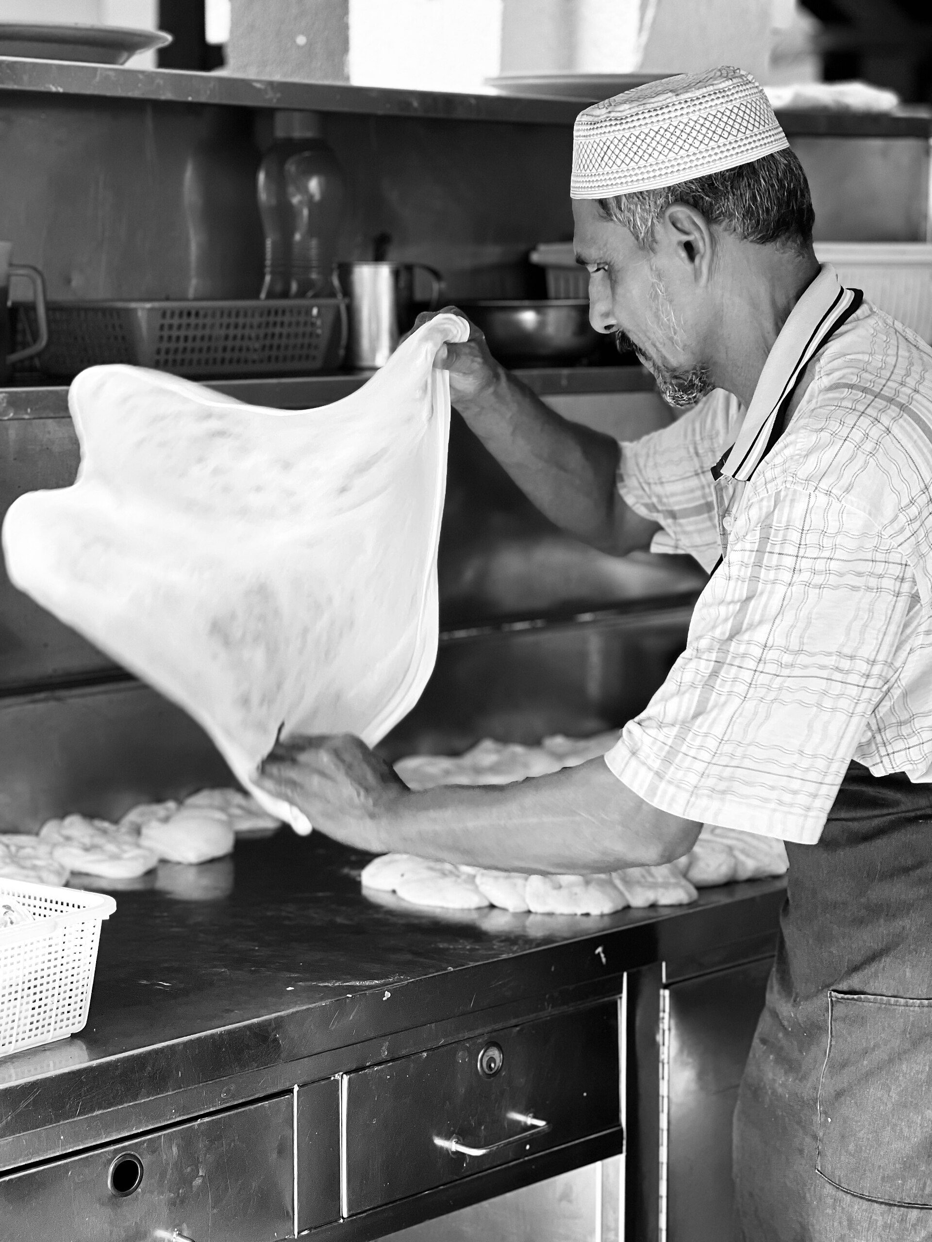 A roti canai maker stretches a sheet of dough in the air over a steel counter.